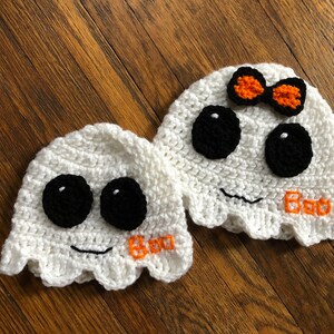 May include: Two white crocheted ghost hats with black eyes and a smiling mouth. One ghost has a small orange bow on top and both say "Boo" in orange yarn.