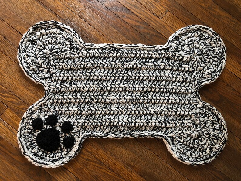 May include: A black and white crocheted dog bone rug with a black paw print in the center.