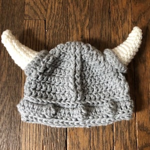 May include: A gray crocheted Viking helmet with two white horns. The handmade hat is designed for a baby or toddler. The helmet is made of soft yarn and sits on a wooden surface.