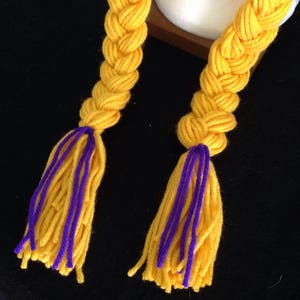 Pu&ograve; includere: Due trecce di filato giallo con accenti di filato viola e nappe. Le trecce sono intrecciate e legate insieme in cima.
