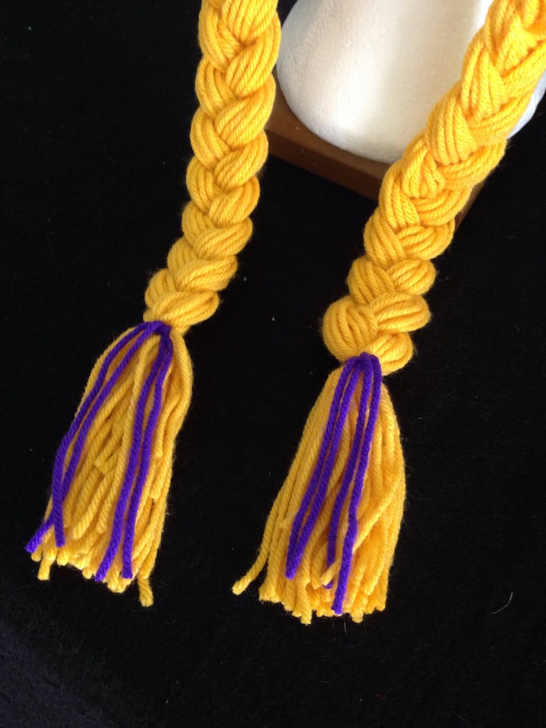 Pu&ograve; includere: Due trecce di filato giallo con accenti di filato viola e nappe. Le trecce sono intrecciate e legate insieme in cima.