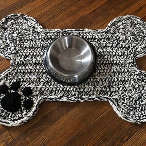 May include: A black and white crocheted dog bone shaped mat with a paw print design and a silver metal food bowl in the center.