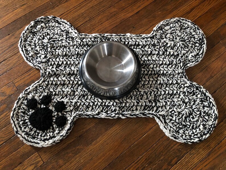 May include: A black and white crocheted dog bone shaped mat with a paw print design and a silver metal food bowl in the center.