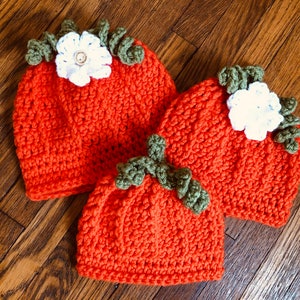 May include: Three orange crochet hats, resembling pumpkins, are arranged on a wooden surface. Each hat features green leaf accents and a white flower with a button center. The hats vary in size, suggesting a set for different ages.