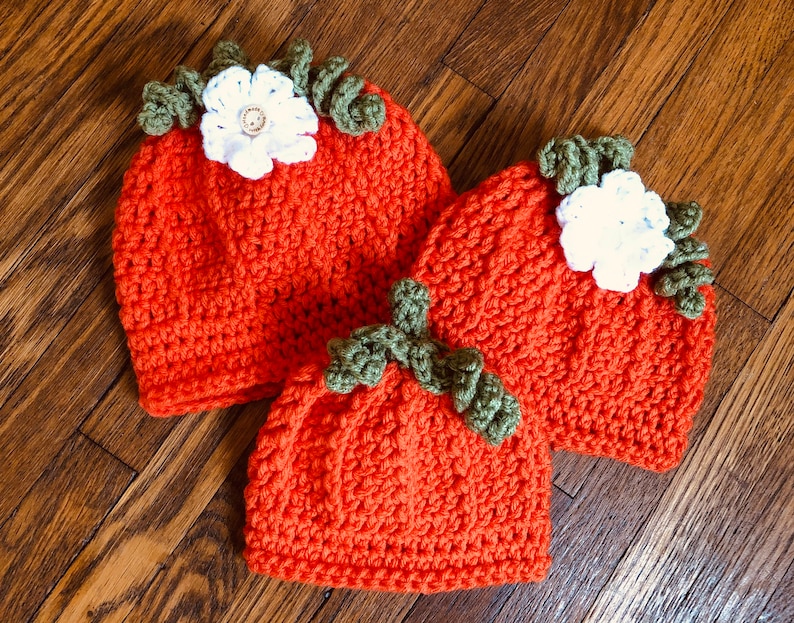May include: Three orange crochet hats, resembling pumpkins, are arranged on a wooden surface. Each hat features green leaf accents and a white flower with a button center. The hats vary in size, suggesting a set for different ages.
