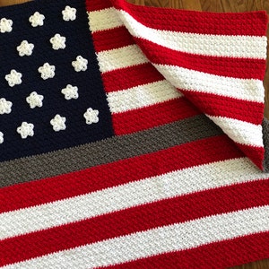 May include: A crocheted blanket in the style of the American flag. The blanket features red and white stripes, a blue field with white star appliques, and a gray stripe. The blanket is folded over, showing the design.