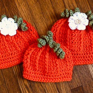 May include: Three handmade crochet pumpkin hats in varying sizes. The hats are orange with green stems and white flower accents. The hats are displayed on a wooden surface.