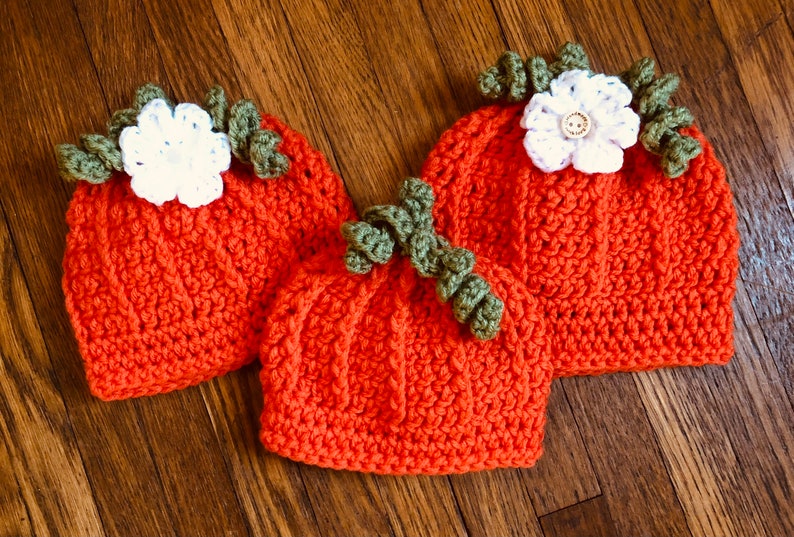 May include: Three handmade crochet pumpkin hats in varying sizes. The hats are orange with green stems and white flower accents. The hats are displayed on a wooden surface.