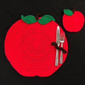 Può includere: Un sottopiatto rosso a forma di mela lavorato a uncinetto con foglie verdi e un gambo marrone. Una forchetta e un coltello sono posizionati sul sottopiatto. Un sottobicchiere rosso a forma di mela lavorato a uncinetto più piccolo è sullo sfondo.