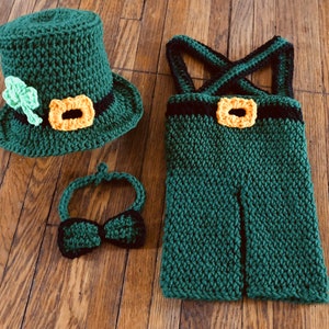 May include: A green crocheted leprechaun costume for a baby, including a hat, overalls, and a bow tie.