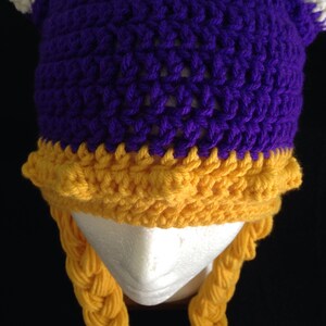 Pu&ograve; includere: Cappello all'uncinetto viola e oro con trecce. Il cappello ha una cima a punta e due corna. Le trecce sono attaccate alla parte inferiore del cappello.