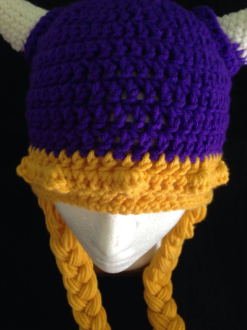 Pu&ograve; includere: Cappello all'uncinetto viola e oro con trecce. Il cappello ha una cima a punta e due corna. Le trecce sono attaccate alla parte inferiore del cappello.