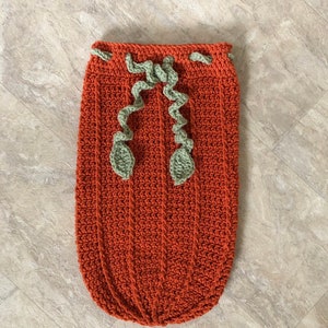 Op de afbeelding: Een gehaakte baby slaapzak in de vorm van een pompoen in oranje met groene gehaakte bladeren.
