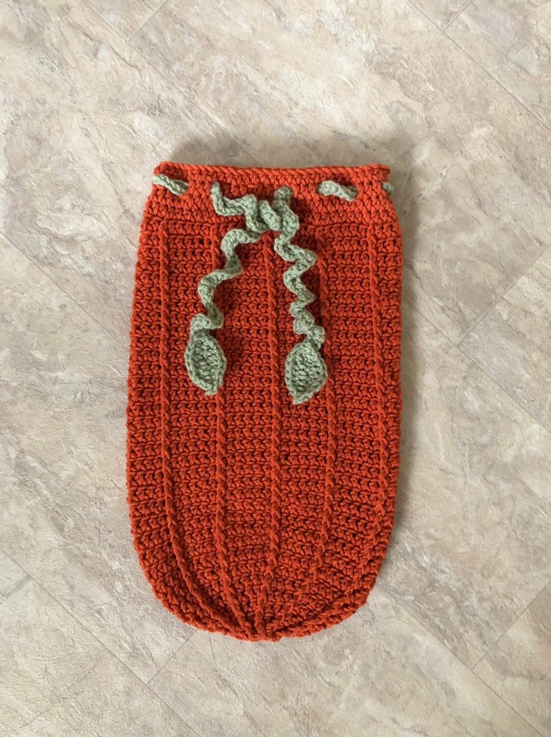 Op de afbeelding: Een gehaakte baby slaapzak in de vorm van een pompoen in oranje met groene gehaakte bladeren.