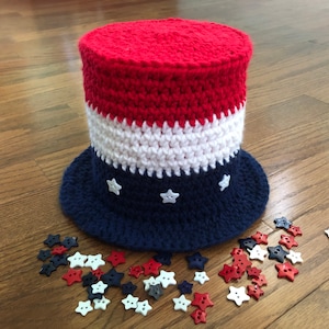 May include: A red, white and blue crocheted hat with a brim and white star buttons. The hat is lying on a wooden surface with a scattering of red, white and blue star buttons.