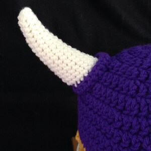 Pu&ograve; includere: Un cappello all'uncinetto viola e oro con un corno all'uncinetto bianco. Il cappello &egrave; realizzato con un punto a trama.