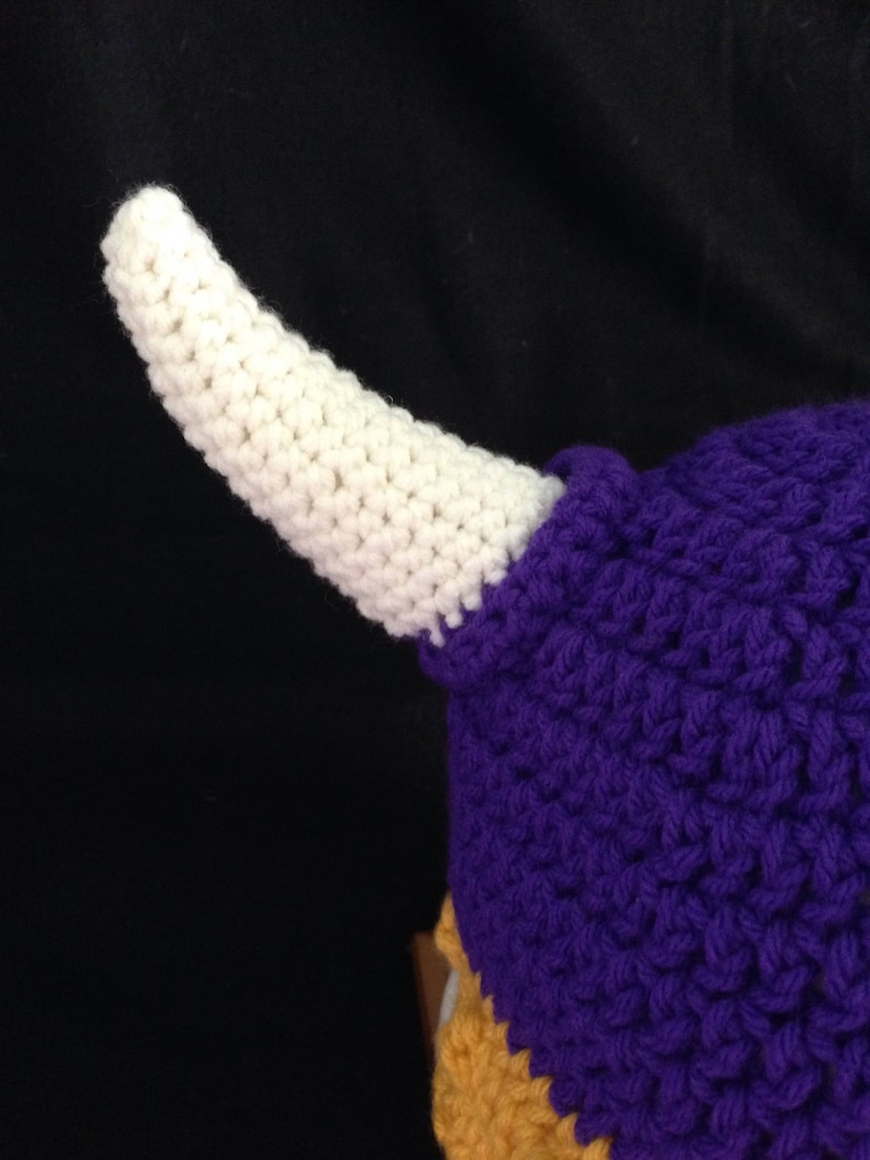 Pu&ograve; includere: Un cappello all'uncinetto viola e oro con un corno all'uncinetto bianco. Il cappello &egrave; realizzato con un punto a trama.