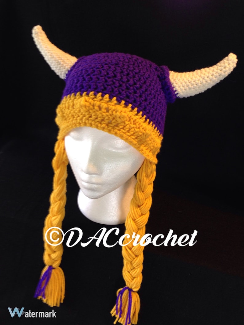 Pu&ograve; includere: Cappello vichingo all'uncinetto viola e oro con corna bianche e trecce gialle. Il cappello &egrave; su una testa di manichino bianca. Il testo "DACcrochet" &egrave; sul cappello.