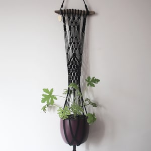 May include: A black macrame plant hanger with a wooden dowel at the top, holding a burgundy pot with green leafy plants. The plant hanger is suspended against a white wall.