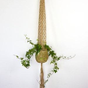 Pode incluir: Um suporte de plantas suspenso em macramê feito de fibra de juta natural. O suporte de plantas suspende um vaso de vidro verde cheio de plantas verdes com folhas. O design em macramê apresenta um anel de madeira na parte superior para pendurar.