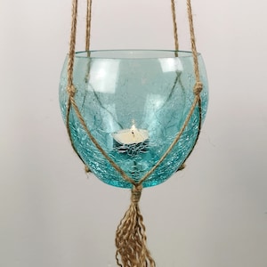 Può includere: Un portacandele in vetro turchese sospeso con una texture screpolata. La ciotola di vetro è sospesa da spago naturale, con una nappa sul fondo. Una candela a lumino accesa è all'interno della ciotola, creando un bagliore caldo.