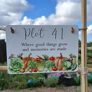 May include: A rectangular white sign reading 'Plot 41' and 'Where good things grow and memories are made'. The sign features illustrations of vegetables and is attached to a bamboo frame. The background shows a garden plot.