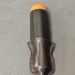 Peut inclure: Un étui à stylo en cuir noir avec des coutures rouges. L'étui a un rabat qui recouvre le haut du stylo. Le stylo est noir avec un dessus orange.