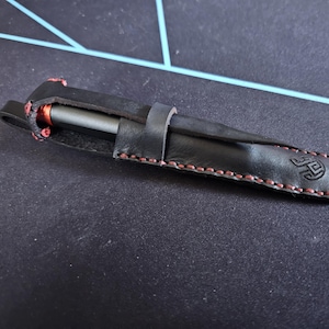 Peut inclure: Un étui à stylo en cuir noir avec des coutures rouges et un petit logo sur le côté. L'étui est fermé et un stylo est à l'intérieur.