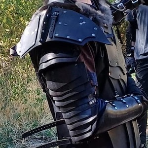 May include: Black leather armor with a layered design, featuring a shoulder plate and a forearm guard. The armor is decorated with metal rivets and has a textured surface.