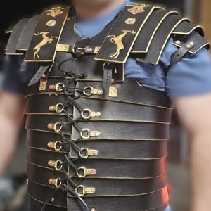 Könnte beinhalten: Schwarzer Leder-Brustpanzer mit goldfarbenen Akzenten und Hirsch-Emblemen. Die Rüstung hat geschichtete Platten, Schulterstücke und einen geschnürten Frontverschluss. Das Design erinnert an historische römische Rüstungen.