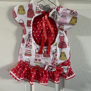 May include: Red and white polka dot apron with a white background featuring a pattern of colorful aprons. The apron has a ruffle trim and a bow at the waist. The text "love the cook" is printed on the apron.