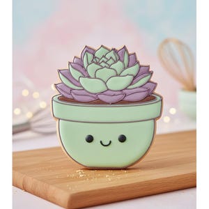 May include: A succulent-shaped cookie with a smiling face, in a light green pot with a brown interior. The succulent has light green leaves with purple tips. The cookie sits on a wooden cutting board.