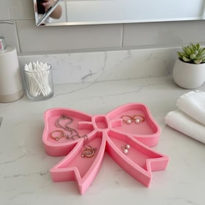 Puede incluir: Una bandeja rosa con forma de lazo para joyas con múltiples compartimentos. La bandeja contiene varias joyas, incluyendo collares, anillos y pendientes. La bandeja está hecha de un material sólido y se coloca sobre una encimera blanca. Una pequeña planta suculenta en una maceta blanca está en el fondo.