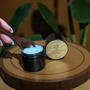 May include: Two small black jars of unscented mineral sunscreen are displayed on a wooden tray. One jar is open, with a wooden spoon scooping out the white cream. The label reads "100% Organic Reef-Safe" and "Golden Trail Skincare".