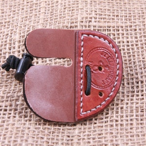 May include: A brown leather archery finger tab with white stitching and a black cord. The tab has a rounded shape with a cut-out section and a black plastic and cord attachment. The item is on a textured, tan background.