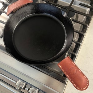 May include: A black cast iron skillet with a leather handle and a leather heat shield. The skillet is on a stainless steel stovetop.