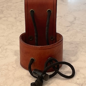 May include: A brown leather holster with a cylindrical base and a rectangular top. Black cord with a plastic adjuster is attached. The holster is designed to hold an item, such as a knife or tool.