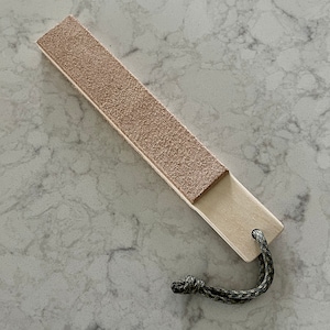 May include: A light brown leather strop with a wooden handle and a green cord attached to the handle.
