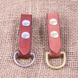 May include: Two leather key fobs with metal rings. The fob on the left has a silver ring and the fob on the right has a gold ring. Both fobs have two silver rivets.