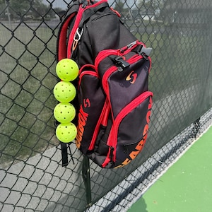 May include: A black and red sports backpack with a bright orange zipper pull. A stack of four yellow pickleball balls are hanging from the backpack.