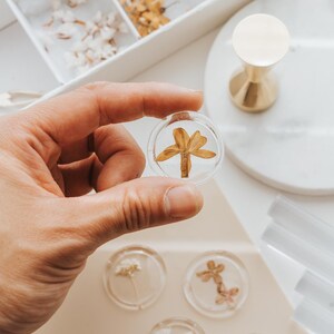 Clear wax seals with dried flowers. One seal, held in a hand, features a dried flower. Other seals with various flowers are visible, alongside a white tray of dried flowers and a gold-colored wax stamp.