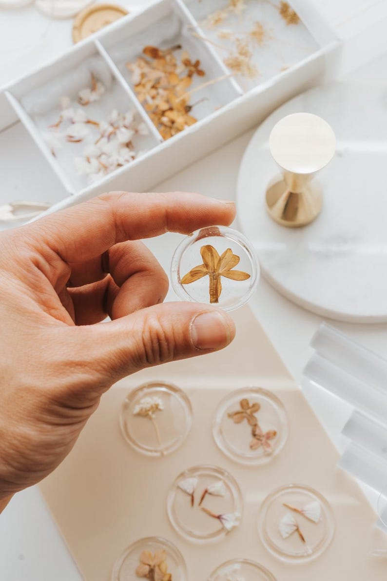 Clear wax seals with dried flowers. One seal, held in a hand, features a dried flower. Other seals with various flowers are visible, alongside a white tray of dried flowers and a gold-colored wax stamp.
