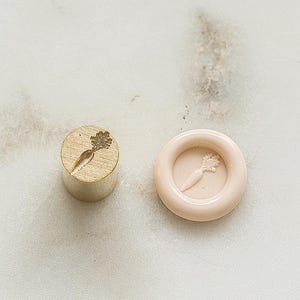 May include: A wooden carrot-shaped stamp and a light pink wax seal with a carrot impression. The stamp is cylindrical and the seal is round with a raised border. The carrot design is detailed.