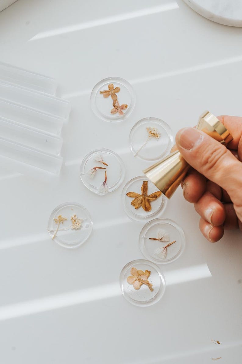 Clear wax seals with dried flowers and a gold-colored wax stamp. The seals are round and contain various dried floral elements. A hand holds the wax stamp, ready to create impressions.