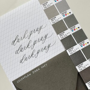 May include: A stationery set featuring a color palette guide and a notebook with the words "dark gray" written in cursive. An envelope with the text "COLORPLAN DARK GREY" is also present. The color guide displays various shades of gray.