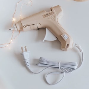 May include: A beige hot glue gun with a white cord and plug. The glue gun has a white trigger and a black switch. The glue gun has the text "milliard" printed on the side.