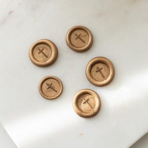 May include: Five gold-coloured wax seals, each featuring a cross design, are displayed on a white marble surface. The circular seals have a raised cross motif. The lighting accentuates the texture and metallic sheen of the seals.