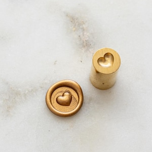 May include: A gold-coloured wax seal stamp and a wax seal with a heart design. The stamp is cylindrical with a heart-shaped impression on the top. The wax seal is round with a heart in the centre. The items are on a white marble surface.