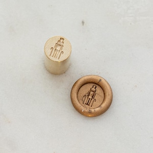May include: Two wax seal stamps. One is a cylindrical wooden stamp with a detailed design of a soldier. The other is a bronze-colored wax seal with the same soldier design. Both are on a white marble surface.