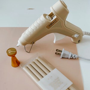May include: A beige hot glue gun with a white plug and cord. There are six beige glue sticks in a clear plastic package with the text "Switch & Wood" and "High Quality Glue Sticks" on the package. There is also a gold metal stamp with a floral design.
